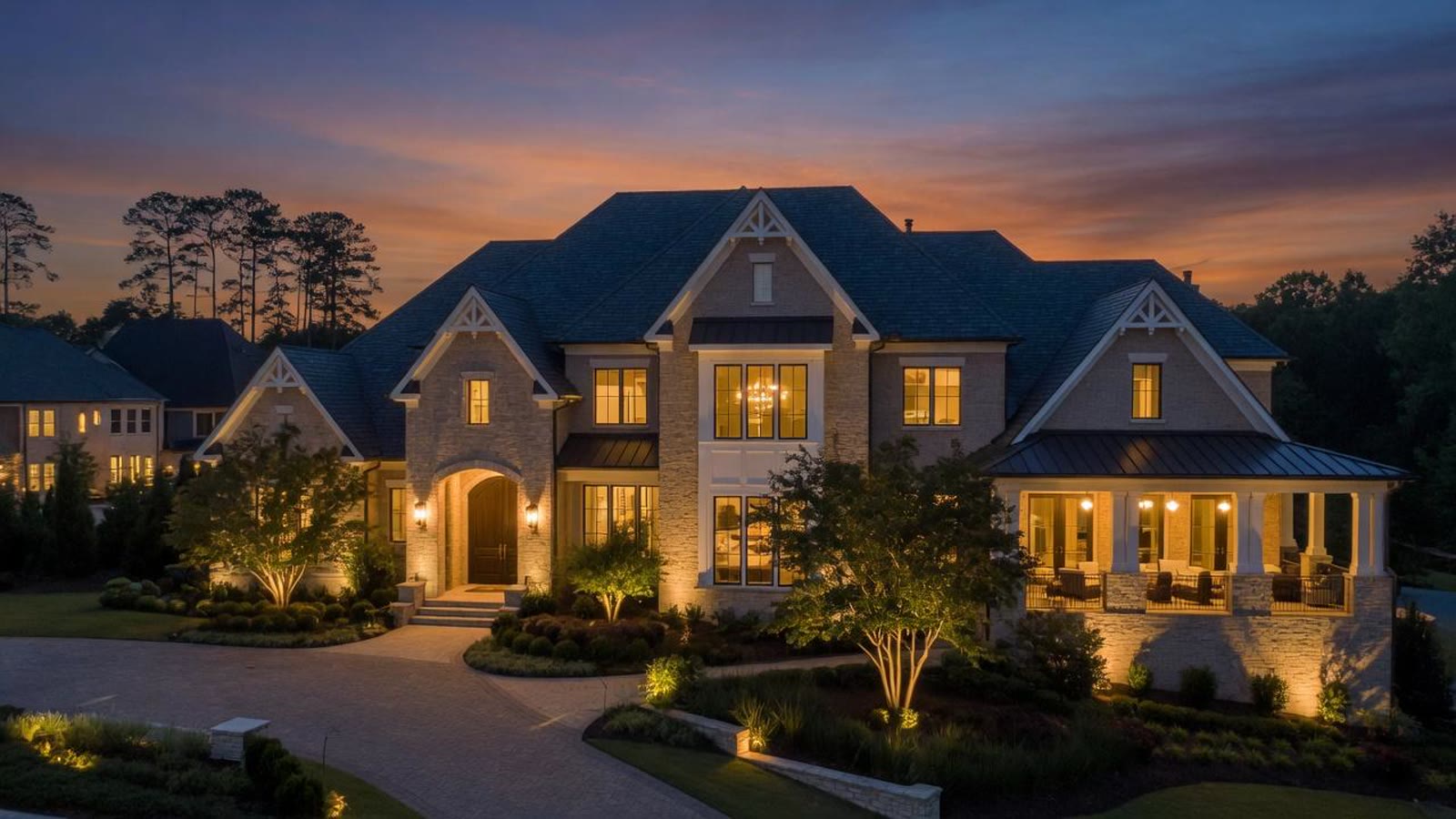 A North Atlanta estate at dusk with full landscape lighting — uplit trees, facade lighting, glowing windows and a lit covered porch against a sunset sky